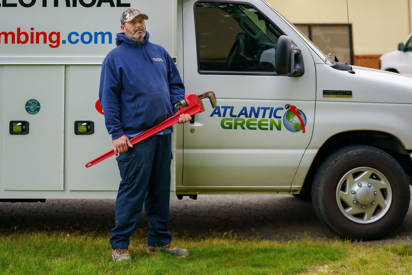A technician holding a red pipe wrench beside an Atlantic Green truck
